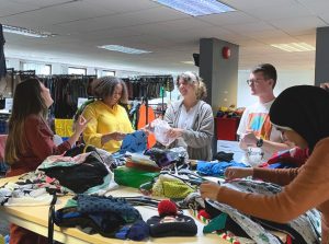 Five people standing around a table are sorting through clothing donations in a bright, spacious room. Shelves of neatly organised garments are in the background.
