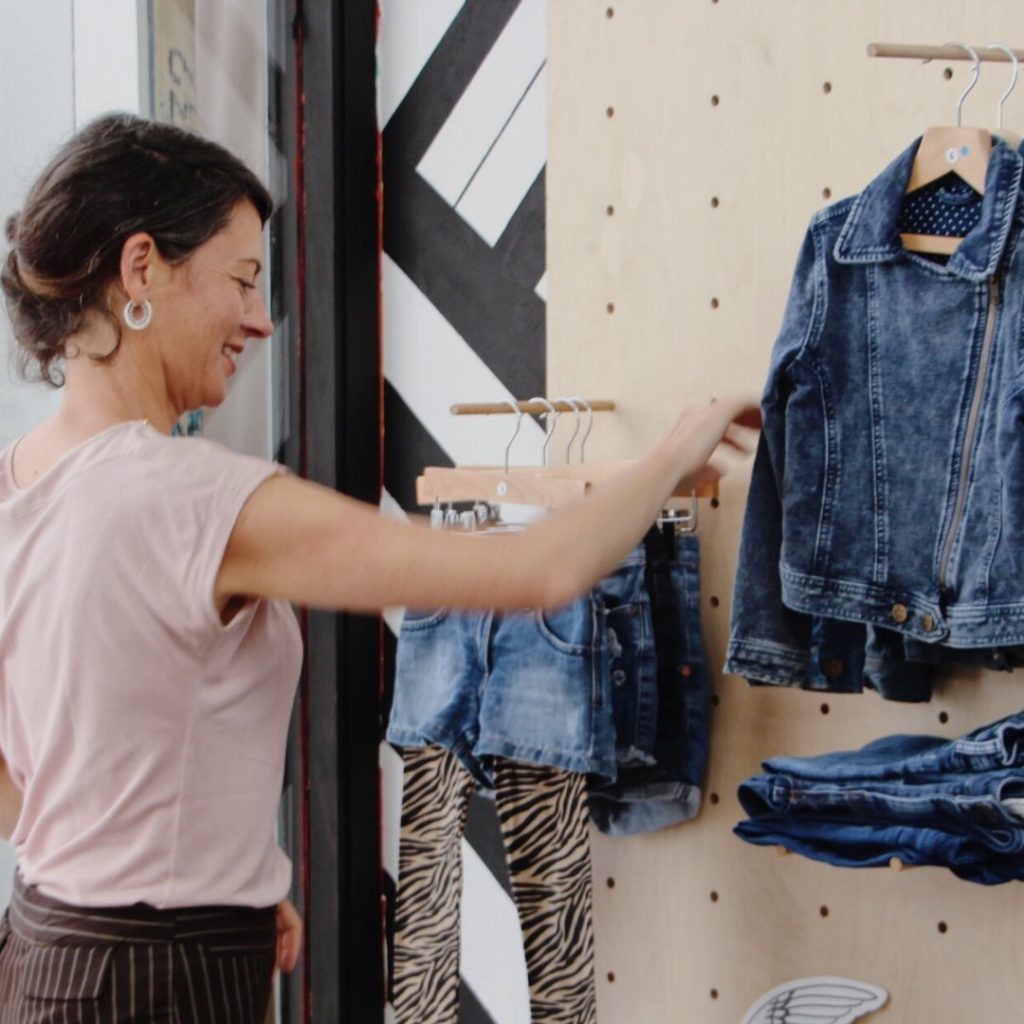 A smiling woman is stood in front of a clothing display featuring denim jackets and shorts. They have their arm raised about to sort through the jackets.